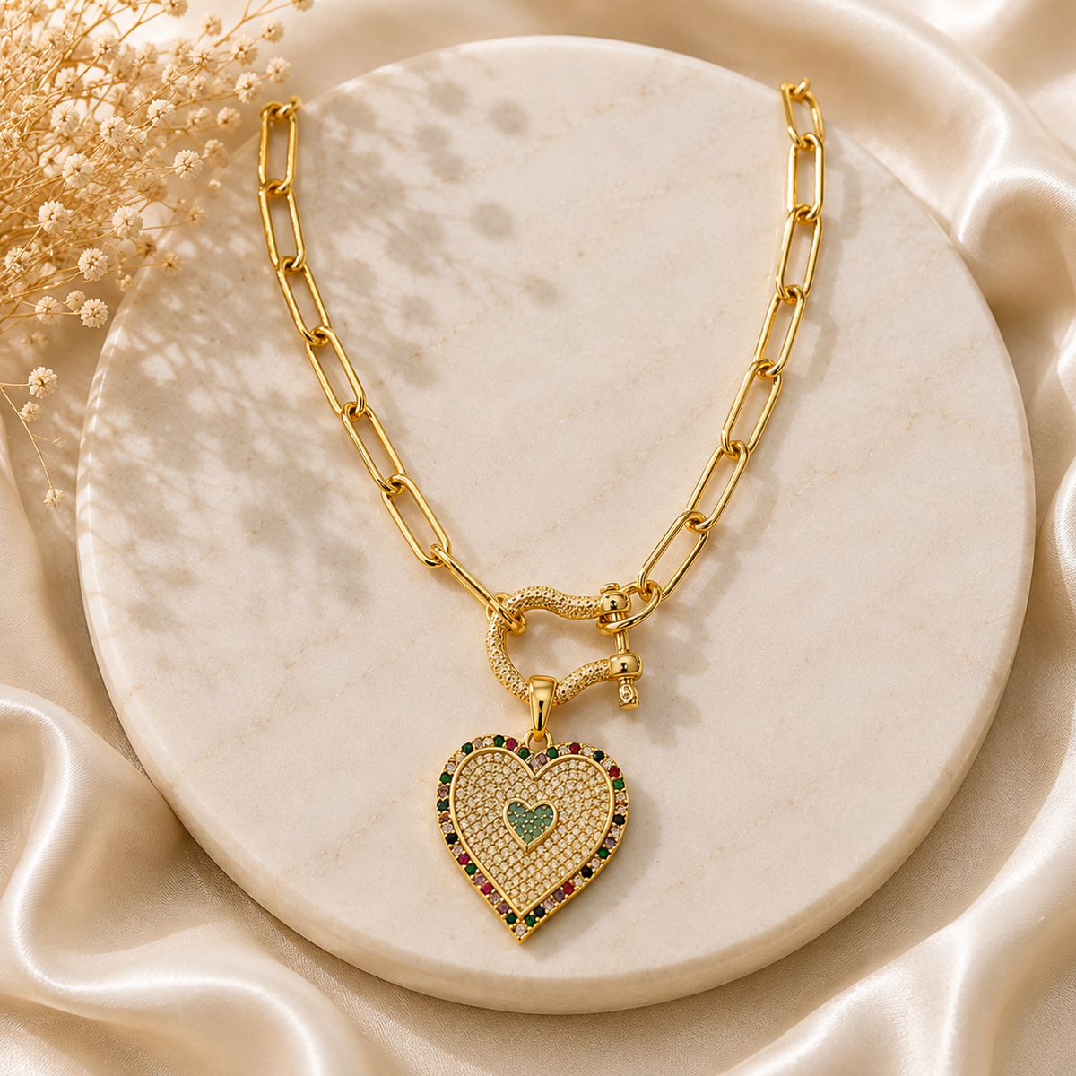 A gold heart pendant necklace with a pavé-set rhinestone border and a small green heart in the center, displayed on a round cream marble slab with satin fabric and dried flowers.
