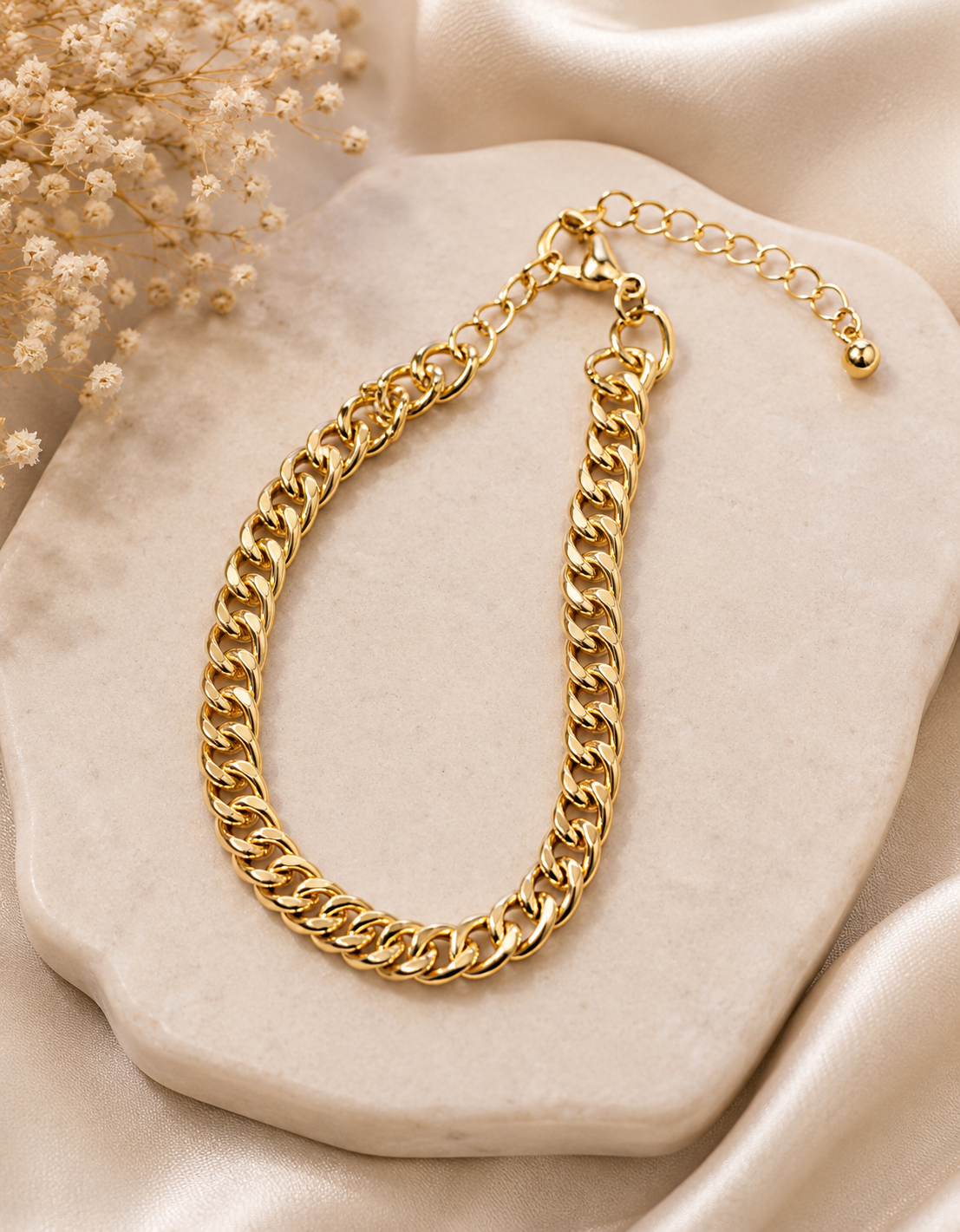 A gold-tone Cuban link chain bracelet with an extender chain and lobster clasp, styled on a beige stone slab with soft satin fabric and dried flowers in the background.